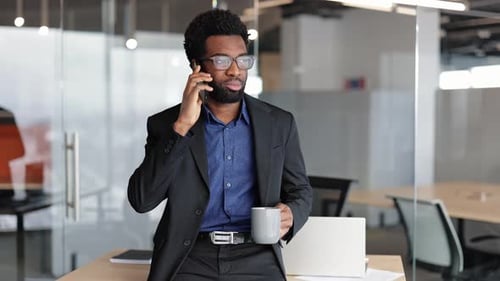 Black Businessman in Suit and Glasses Drinks Coffee and Talks on Phone in Modern Office