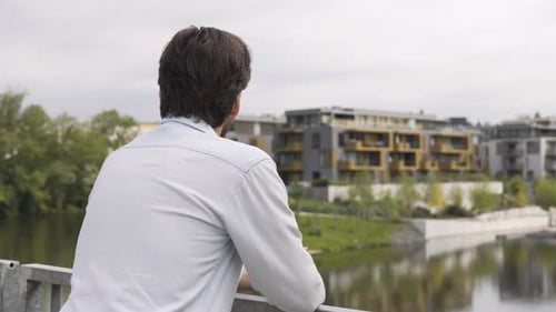 A Man Looks Into a Distance at a Cityscape and a River As He Stands on a Bridge
