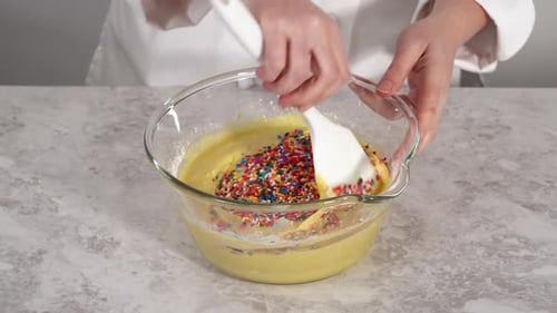 Zusammenfalten von Regenbogenstreuseln zu einem Kuchenteig in einer Glas-Rührschüssel, um Funfettti Bundt-Kuchen zu backen