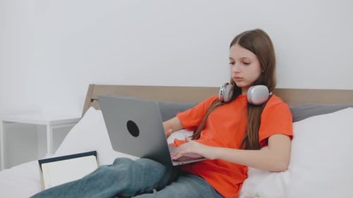Teenager Studies with a Laptop While Wearing Headphones in a Cozy Bedroom Setting During the