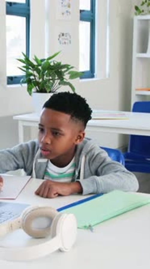 Vertical video: Glancing at laptop, student in gray hoodie writing in notebook for class at desk