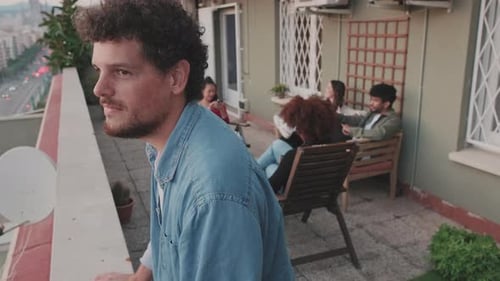 Guy looks at the city standing on the balcony, his friends are chatting in the background