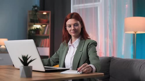 Remote Work Young Woman with Glasses Makes Notes in a Notepad While Sitting at a Computer in Home