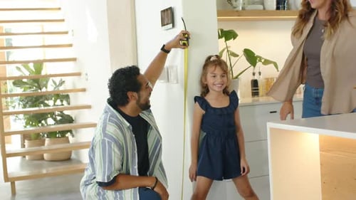 Pai de família ajoelhado estendendo fita amarela, medindo a altura da menina contra a parede na cozinha