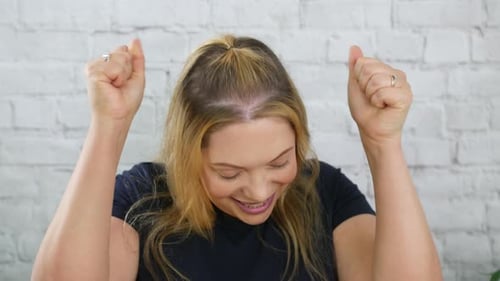 Uma mulher loira feliz comemorando a felicidade sorrindo e vencendo. Dança da vitória contra uma parede de tijolos brancos