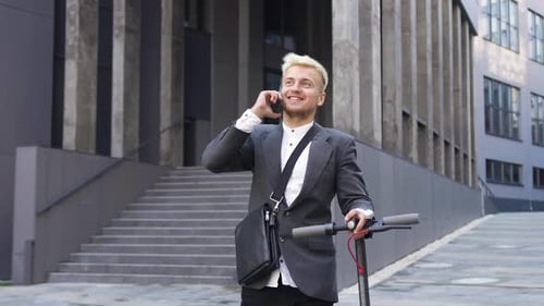 Man in stylish clothes walking with e-scooter near city building and talking on mobile