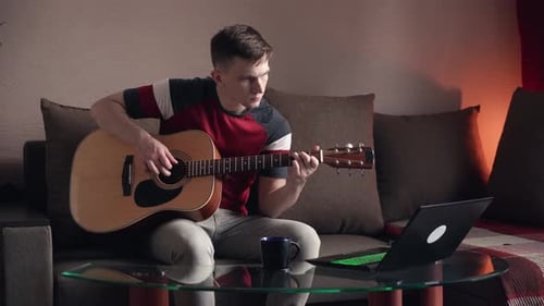 Young Adult Plays Guitar at Home with Laptop