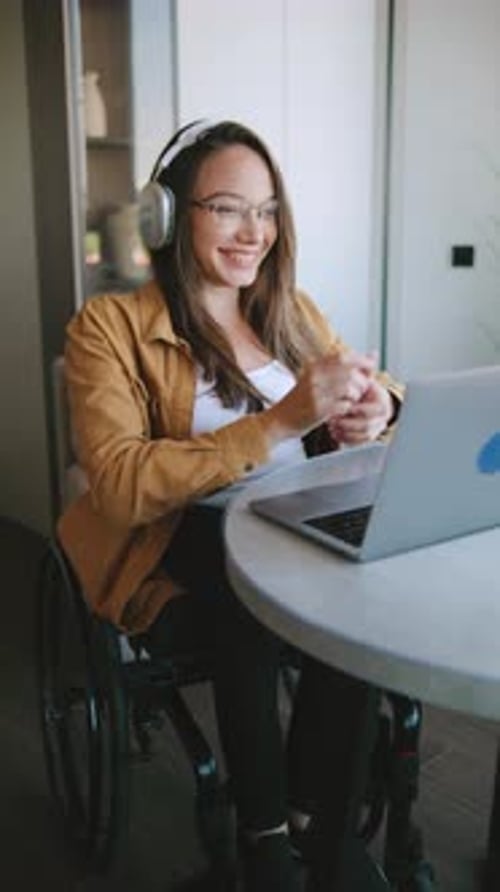 Una mujer en silla de ruedas usa una computadora portátil para trabajar a distancia mientras usa auriculares en un espacio interior moderno con
