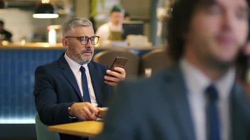 Middle Aged Businessman Drinking Coffee and Browsing Smartphone in Cafe