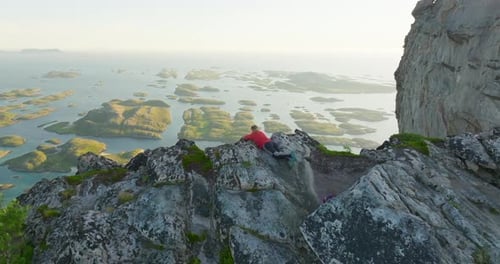 Female hiker on cliff edge overlook scenic Norway islands in midnight sun; drone