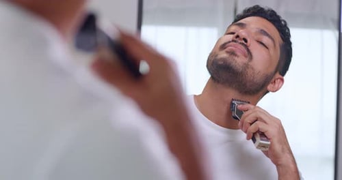 A man is using electric shaver to trim beard while standing in front of mirror.