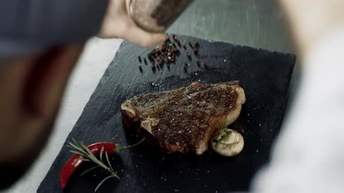 Chef hands peppering grilled steak on stone cutting board at restaurant kitchen