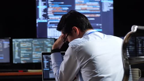 Programador que tiene dolor de cabeza mientras escribe código con una computadora portátil usando varios monitores que muestran la base de datos