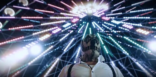 Figure in Mask Standing Before Lit Ferris Wheel