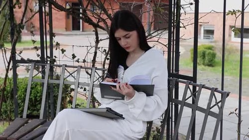 62 1A female student sits on a park bench and studies using a tablet and takes notes.