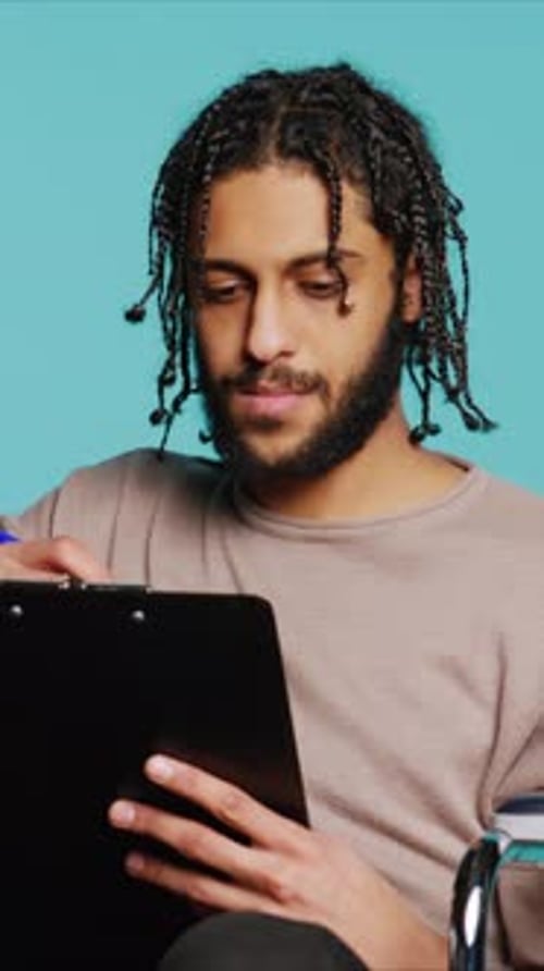 Vertical Video Man in Wheelchair Writing on Clipboard with Pen Studio Background