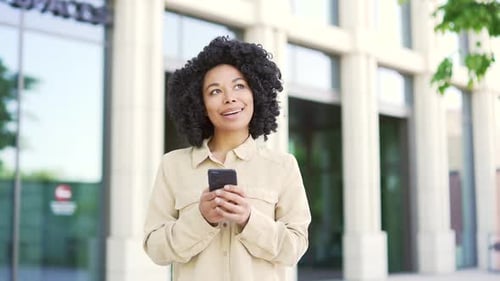 Young african american female student is using smartphone while walking on campus space near