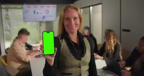 Smiling businesswoman holding smartphone with green screen toward camera during office