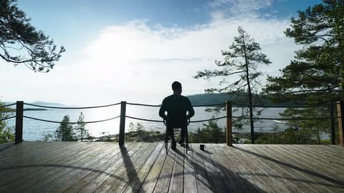 Silhouette Of Tourist Sitting On Balcony Of Luxury Eco Hotel And Admiring Landscape With Calm Lake