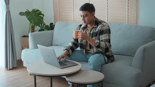 Young Man Uses Laptop While Drinking Coffee At Home