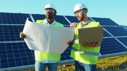 Young Two Engineers Use Laptop and Project Plan on the Solar Farm in Special Uniform Standing