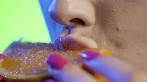 Mulher comendo uma fatia de laranja com luz azul sobre fundo verde em câmera lenta