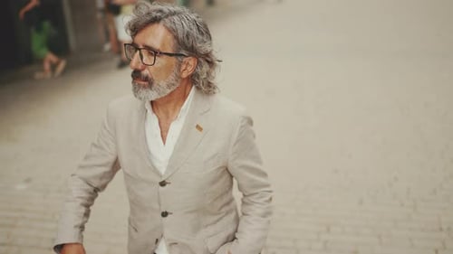 Mature businessman wearing gray jacket walks down the street past modern buildings