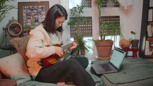 Woman Plays Ukulele While Looking at Laptop Indoors