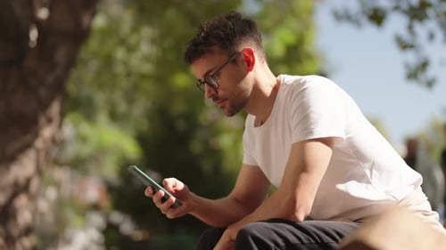 Young Man with Glasses Using Smartphone on Park Bench