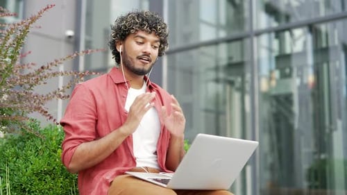 Young businessman talking on a video call using a laptop while sitting on a bench near office