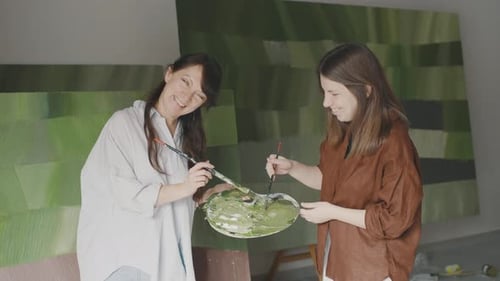 Two women joyfully create abstract art together in a bright studio filled with colorful paintings