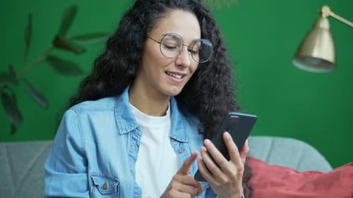 Porträt eines lächelnden brünetten Mädchens mit Brille und Smartphone, das scrollt und soziale Medien schaut