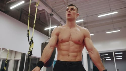 A Young Muscular Man with a Naked Torso Recovers His Breath After a Training in the Gym and Rotates