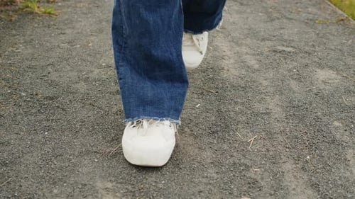 Junge Frau in blauen Jeans und weißen Turnschuhen läuft an sonnigen Tagen auf einer Straße zwischen grünem Gras