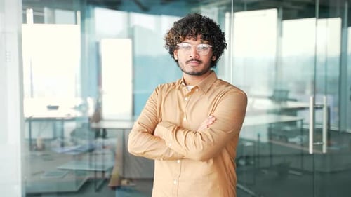 Portrait of young successful handsome man in glasses with crossed arms standing in modern office.
