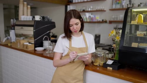 Portrait of a Happy Girl Waitress in a Yellow Apron and White Tshirt Who Makes Notes in Her Notebook