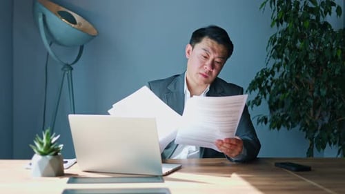 Fatigued Manager Reviewing Documents While Sitting at Desktop with Laptop