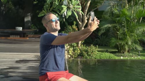 Handsome man taking smartphone photo by luxury swimming pool on summer vacation