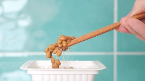 Wooden chopsticks holding Japanese natto beans against background of mint tiles, side view.