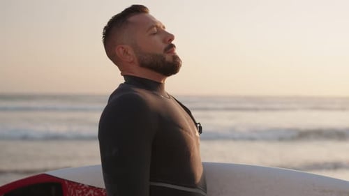 Surfer Stands on Beach at Sunset with Surfboard Gazing at Ocean Waves