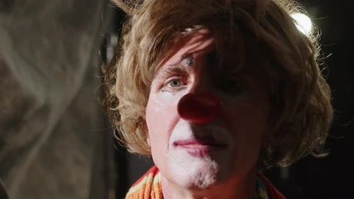 Pensive Clown in Messy Wig and Red Nose Sitting in Circus Backstage Area