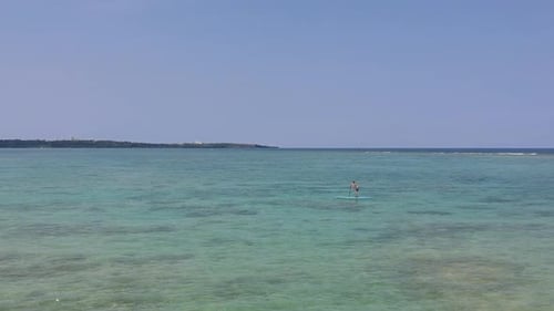 Drone shot of young boy out in the middle of a tropical sea paddling SUP. Boy waves to drone when he