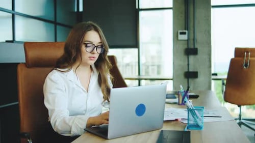 Young Woman Working Intently at a Laptop in a Modern Office with Large Windows on a Sunny Day