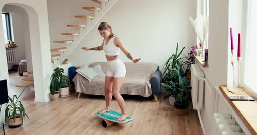 A Blonde Girl in a Bright Room with Plants is Trying to Stand on a Special Device to Improve Balance