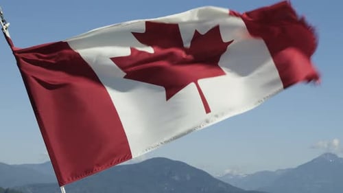 The Canadian flag waving in the wind against a blue sky with green mountains in the distance.