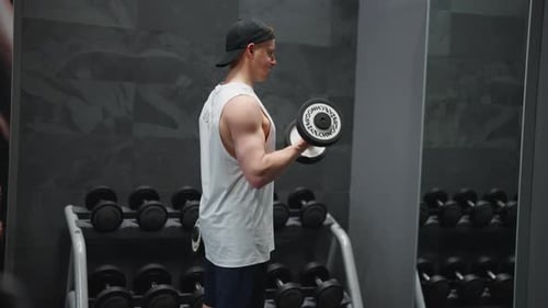 Muscular male do standing dumbbell biceps curl in front of gym mirror