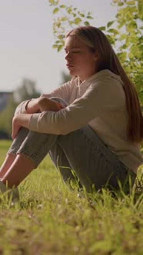 Thoughtful Woman Sitting on Grass in Sunlight with Urban Stadium Background