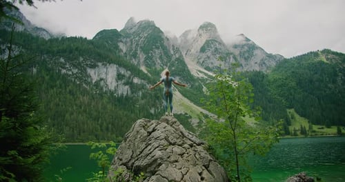 Woman stands on a rocky peak with arms raised, gazing at the towering mountains and lush greenery su