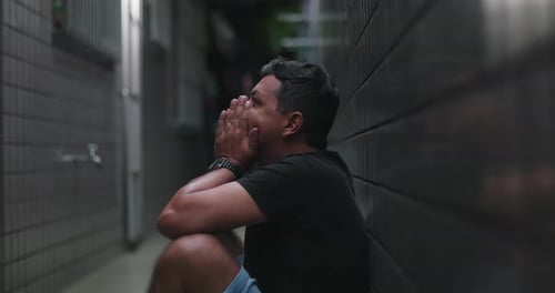 Man sitting with hands covering head in a narrow alley, overwhelmed by despair,