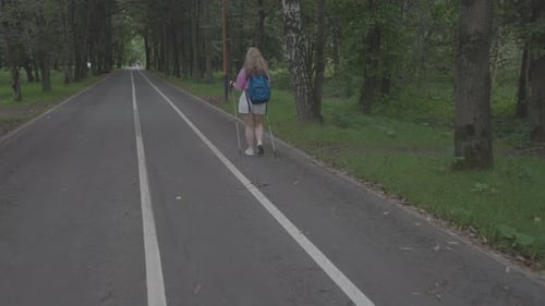 A shot from the back, a girl walking on an asphalt road in a forest, using walking sticks, surrounde